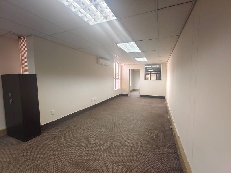 A clean, vacant office space featuring a suspended ceiling with recessed lighting, wall-mounted air conditioning, and neutral carpet flooring.