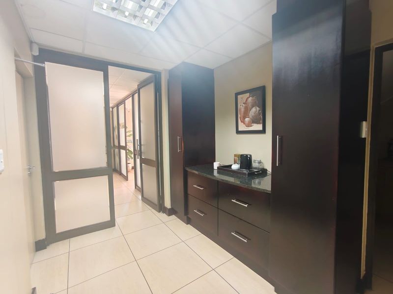 A compact kitchenette area featuring dark wood built-in cabinetry, stone countertops, and tiled flooring, connected to an office hallway via glass partition doors.