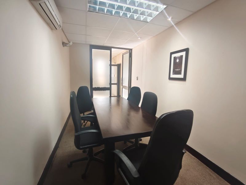 A functional office meeting room featuring a suspended ceiling, recessed lighting, wall-mounted air conditioning, and carpeted flooring with a glass-paneled entrance.