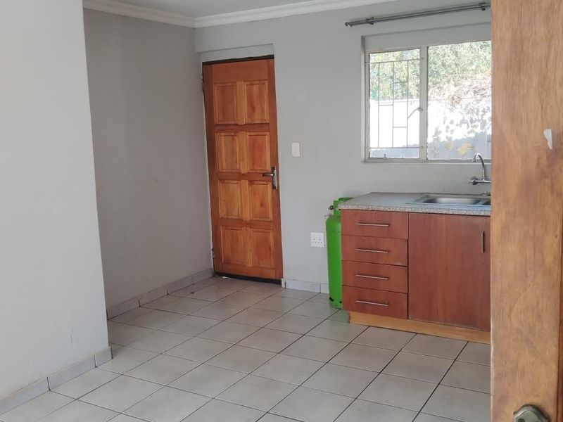 A compact, empty kitchen area featuring tiled flooring, basic wooden cabinetry with a stainless steel sink, and a window with security bars.
