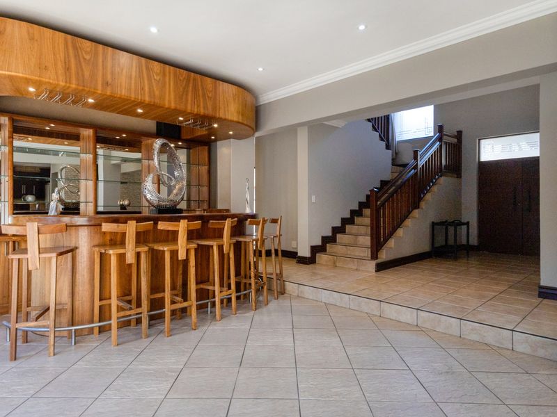 A well-appointed home bar area featuring a custom wooden bar counter, mirrored back, and tiled flooring, adjacent to a staircase and entrance.