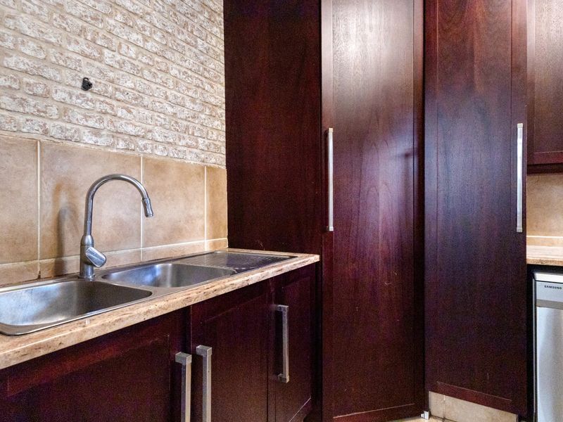 A functional scullery area featuring dark wood cabinetry, a double stainless steel sink, stone-look countertops, and a decorative exposed brick feature wall.