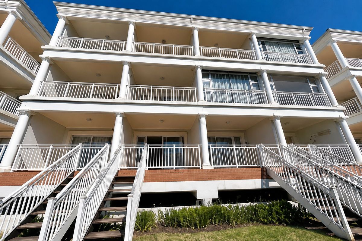A low-angle view of a multi-story apartment building featuring stacked balconies with classical columns, metal railings, and external staircases leading to the ground level.