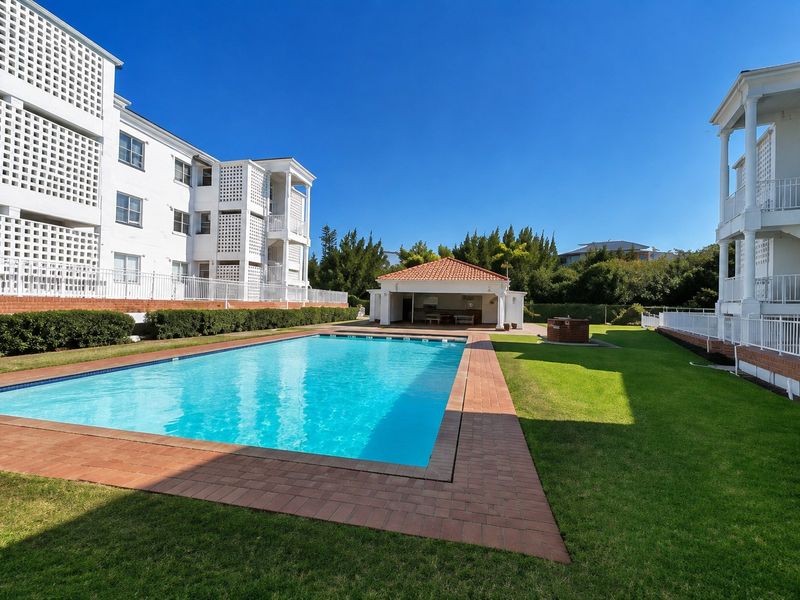 A well-maintained communal swimming pool area featuring brick paving, a manicured lawn, and a covered clubhouse facility within the apartment complex.