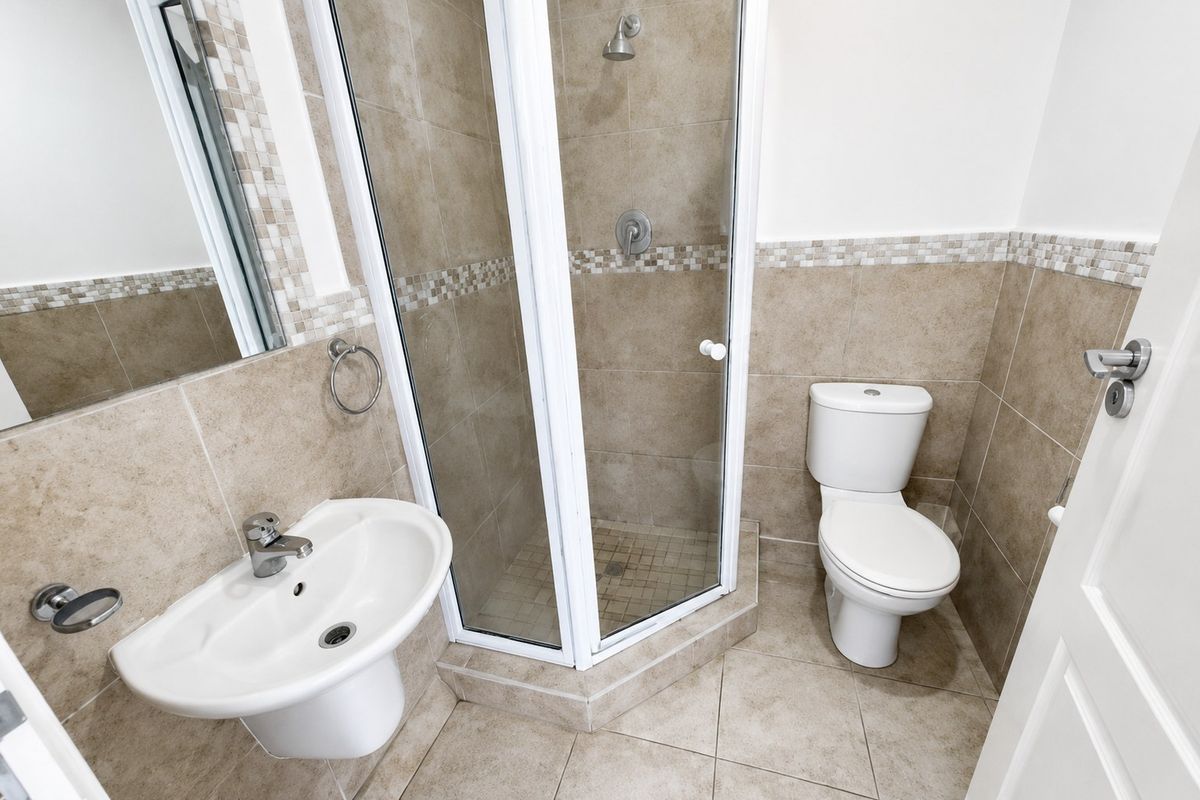 A clean, functional bathroom featuring a corner shower enclosure, pedestal basin, and toilet with neutral tiled finishes.