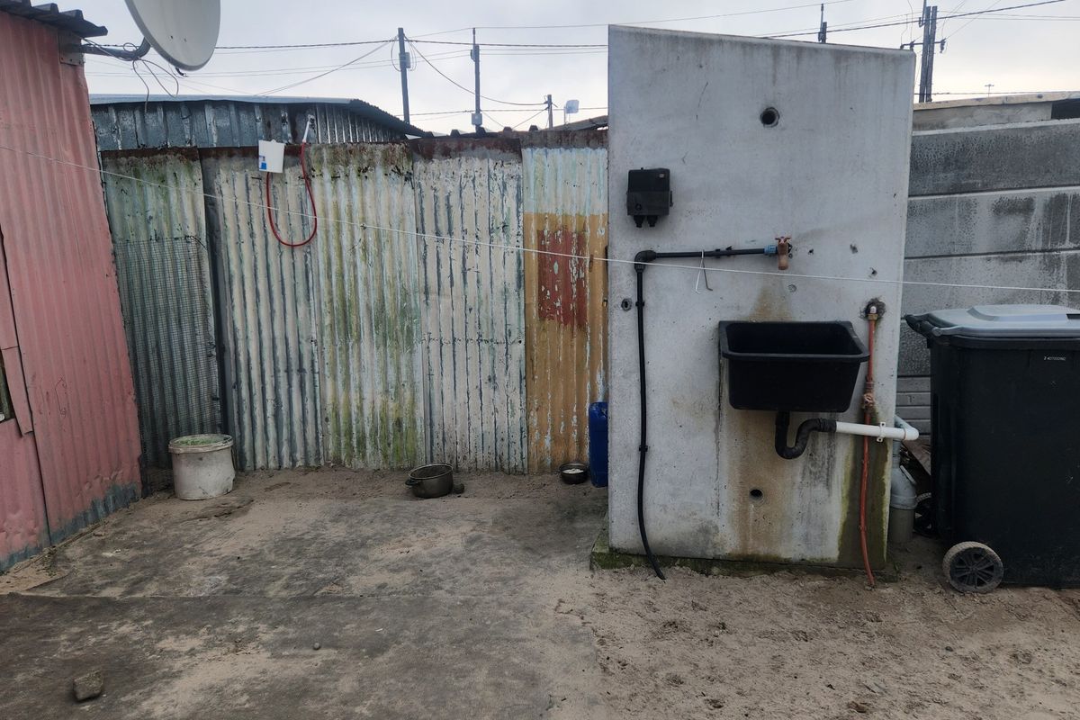 An outdoor utility area featuring a concrete pillar with a mounted sink, plumbing connections, and corrugated metal boundary fencing.