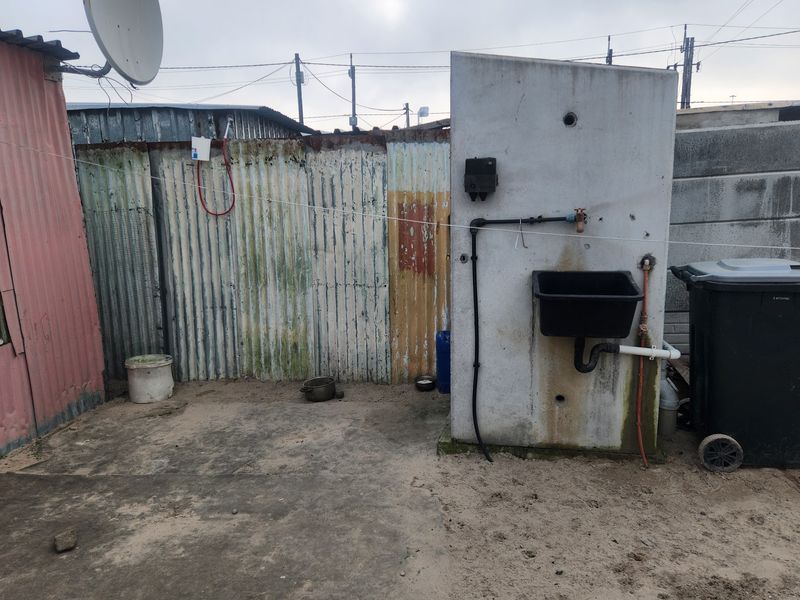An outdoor utility area featuring a concrete pillar with a mounted sink, plumbing connections, and corrugated metal boundary fencing.