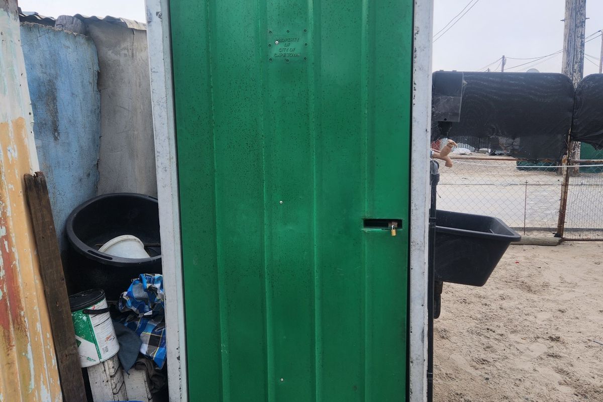 A standalone concrete utility structure with a green metal door, located on a sandy residential plot.
