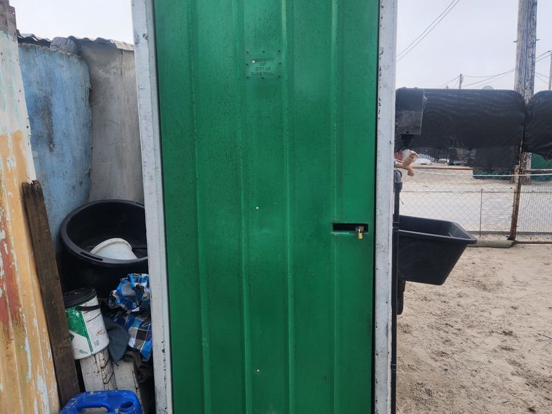 A standalone concrete utility structure with a green metal door, located on a sandy residential plot.