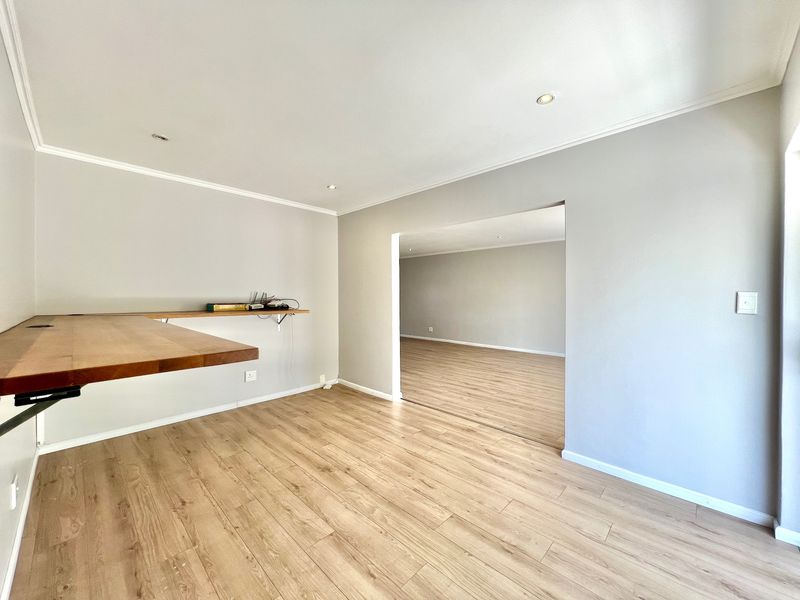 An empty, well-lit living area featuring modern laminate flooring, recessed downlights, and a built-in wooden desk, leading into an adjacent room.