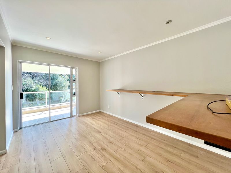 A bright, empty office space featuring modern laminate flooring, recessed lighting, and a built-in wooden desk with access to a private balcony.