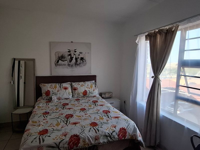 A functional bedroom featuring built-in cupboards, tiled flooring, and a large window with security bars, offering natural light and a view of the surroundings.