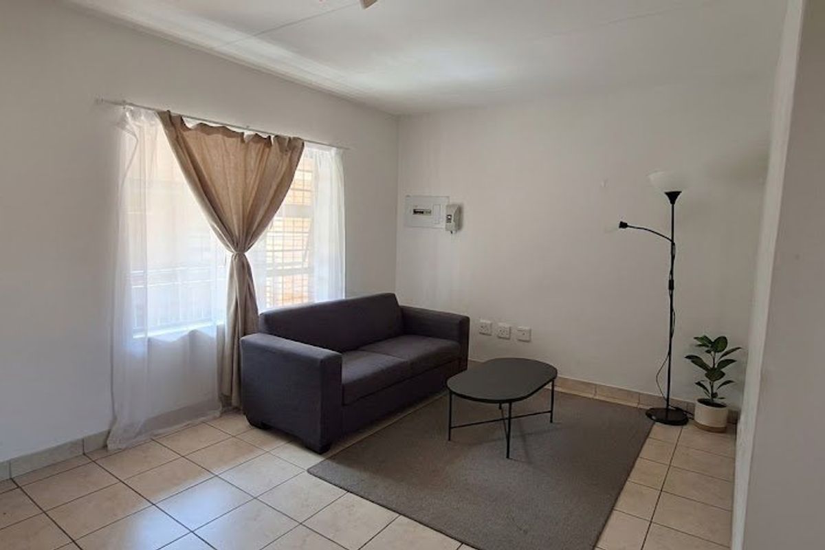 A simple living room featuring neutral tiled flooring and a wall-mounted electrical distribution board, offering a clean and functional space.
