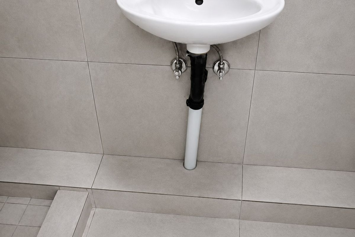A minimalist bathroom featuring a wall-mounted basin with exposed plumbing and neutral ceramic tiling throughout.