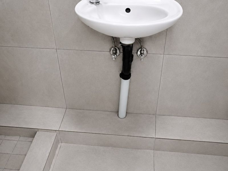 A minimalist bathroom featuring a wall-mounted basin with exposed plumbing and neutral ceramic tiling throughout.