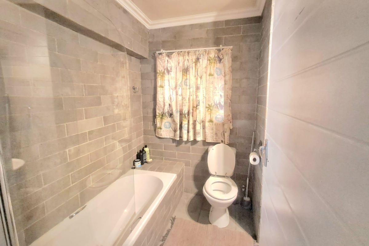 A functional bathroom featuring grey subway-style wall tiles, a built-in bathtub with a glass screen, and a standard toilet.