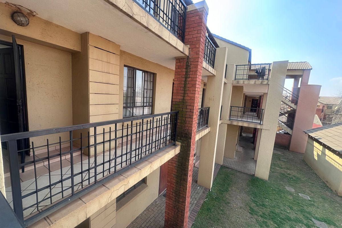 Exterior view of an apartment complex featuring multi-level balconies with metal railings, brick pillars, and paved ground level access.