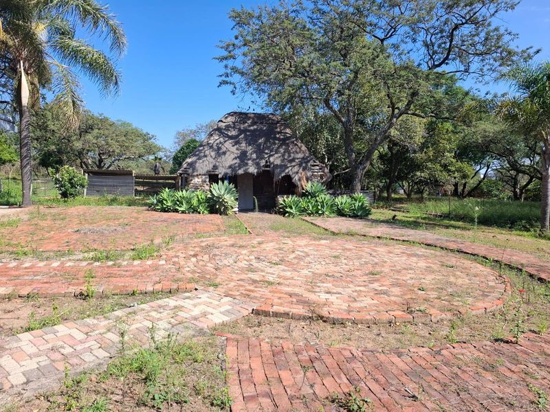 A rustic farm structure featuring a traditional thatched roof and extensive brick-paved courtyard area surrounded by mature trees and garden landscaping.