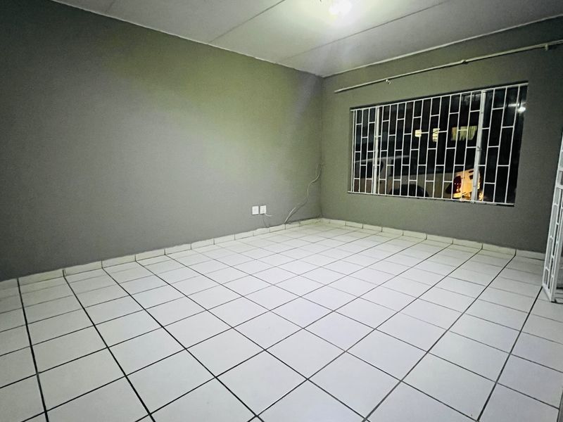 An empty living room featuring neutral grey walls, white tiled flooring, and a window fitted with security bars.