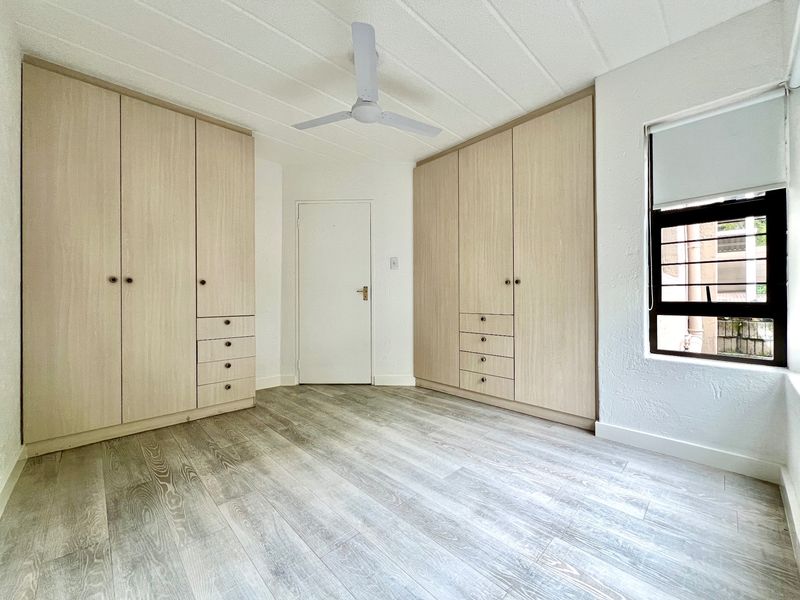A bright, empty bedroom featuring extensive built-in light wood cabinetry, modern laminate flooring, and a ceiling fan.