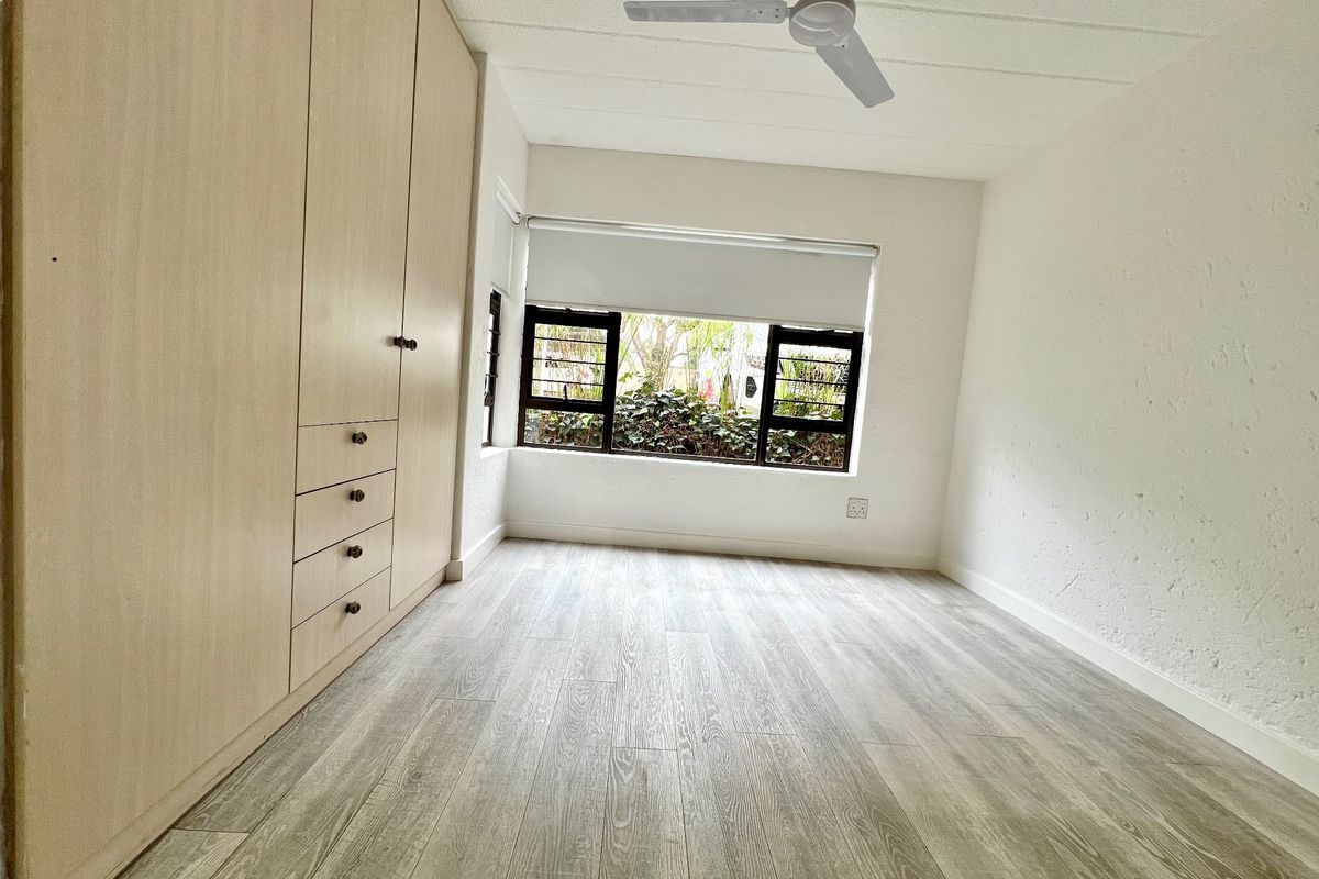 A bright, empty bedroom featuring light wood-tone built-in cupboards, modern laminate flooring, a ceiling fan, and large windows overlooking greenery.
