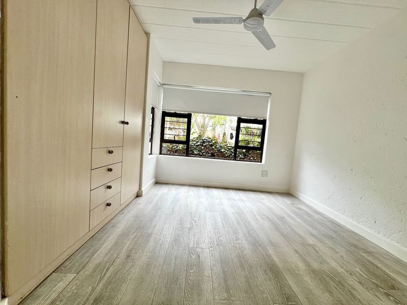 A bright, empty bedroom featuring light wood-tone built-in cupboards, modern laminate flooring, a ceiling fan, and large windows overlooking greenery.
