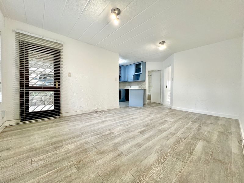 An open-plan living area featuring light-toned laminate flooring, a glass panel door with blinds, and a view into a compact kitchen with blue cabinetry.