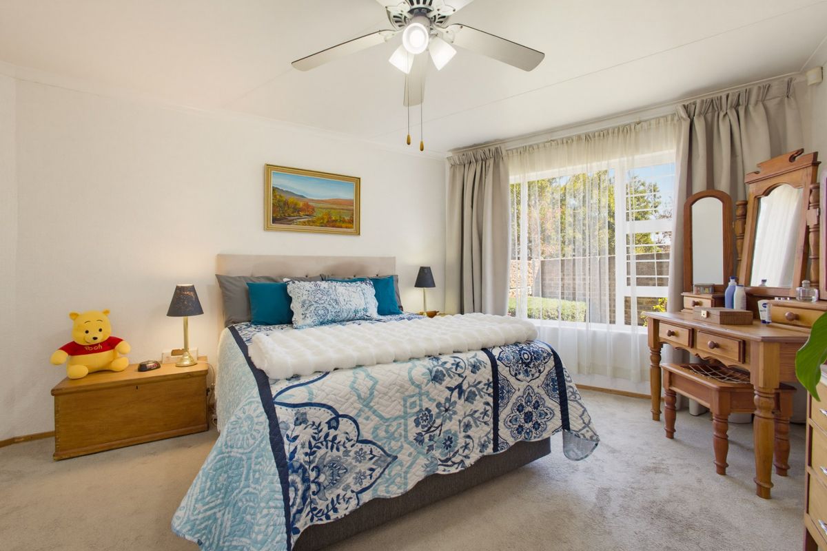 A well-lit bedroom featuring a ceiling fan, large window with natural light, and neutral carpeting, offering a comfortable and functional living space.