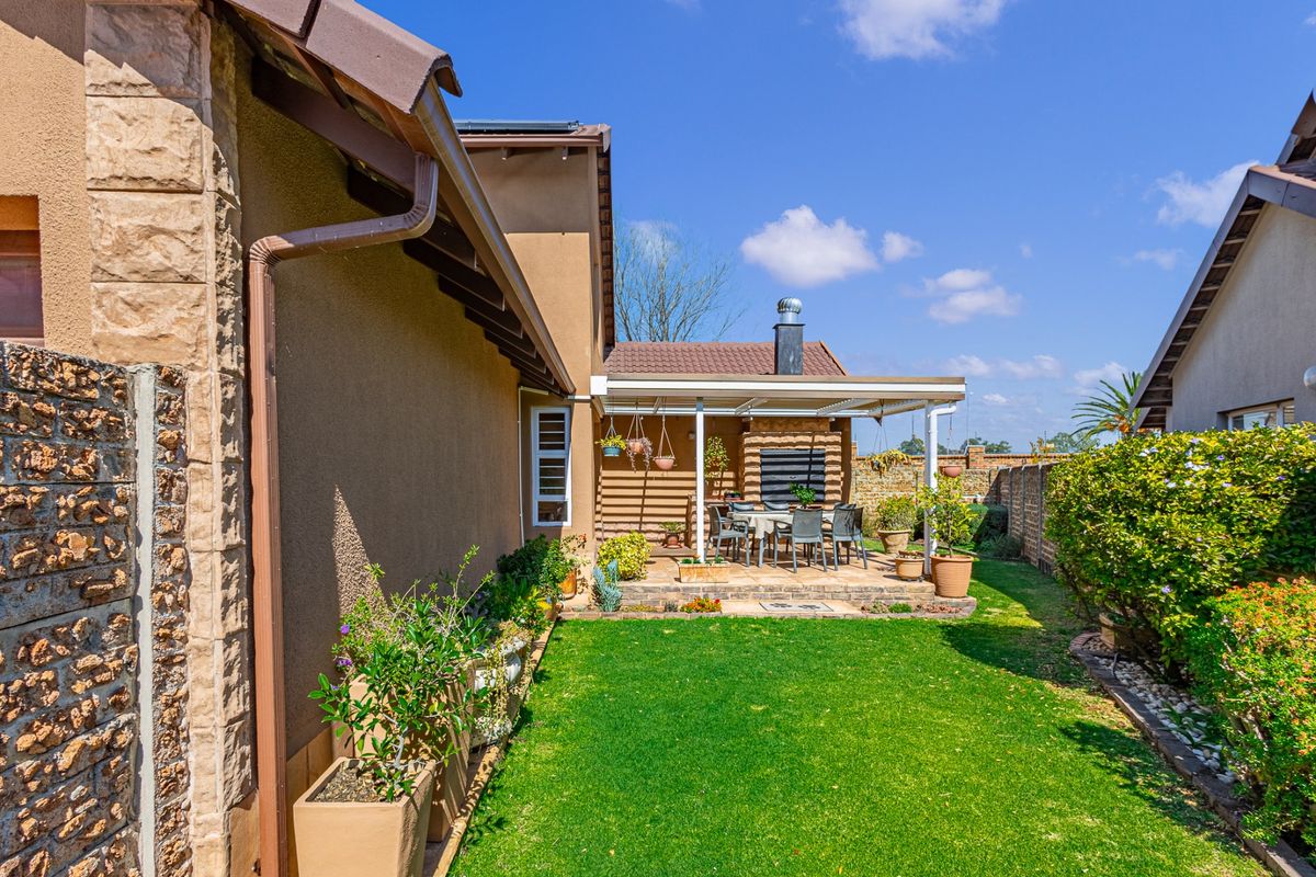 Rear view of the property featuring a well-maintained garden, a covered patio area with a built-in braai, and stone-clad exterior detailing.