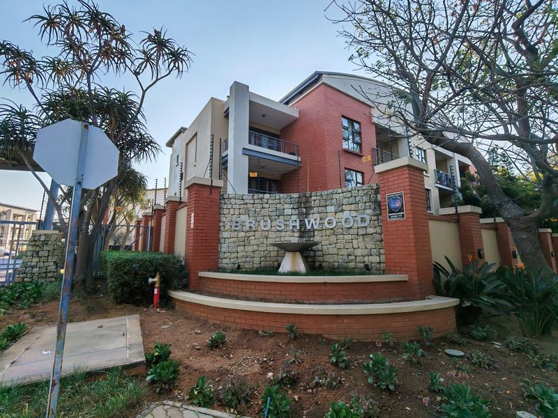 Front entrance view of the Brushwood apartment complex, featuring brick and stone facade architecture with integrated security fencing and landscaped garden beds.