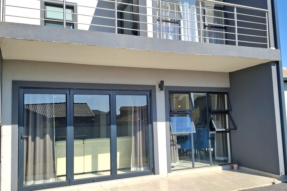 Rear view of a modern double-storey house featuring a tiled patio, aluminium stacker doors, and an upper-level balcony with stainless steel railings.