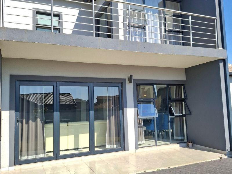 Rear view of a modern double-storey house featuring a tiled patio, aluminium stacker doors, and an upper-level balcony with stainless steel railings.