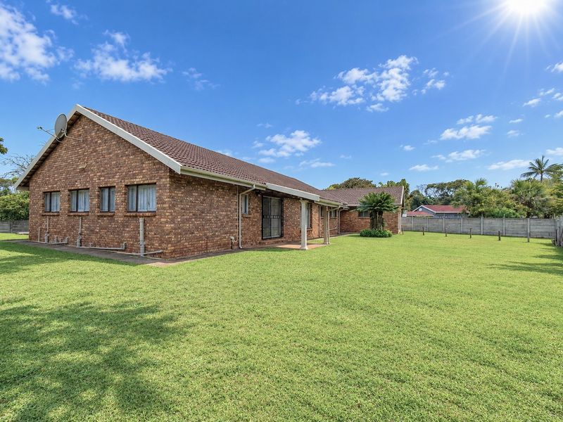 Spacious rear exterior view of a face brick house featuring a large lawn, covered patio area, and secure boundary walling.