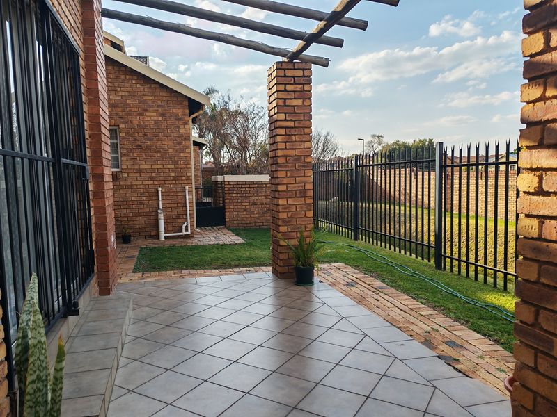 A covered brick patio area featuring tiled flooring, brick pillars, and a pergola, overlooking a private garden space with secure perimeter fencing.