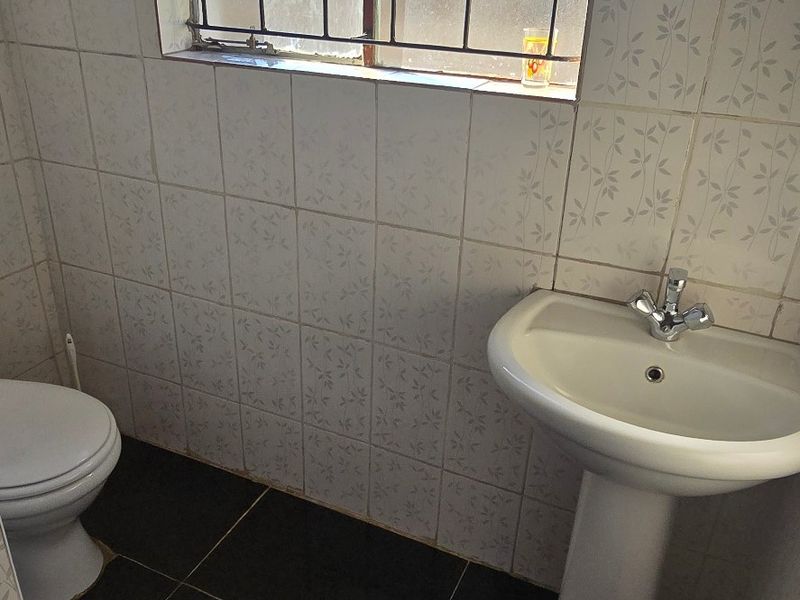 A compact bathroom featuring a pedestal washbasin, toilet, dark floor tiles, and light patterned wall tiles with security bars on the window.