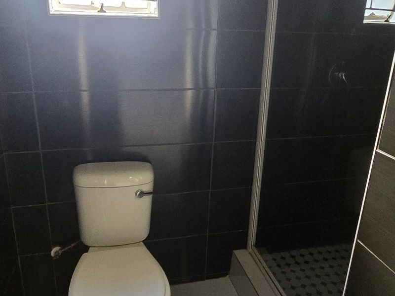 A functional bathroom featuring dark wall tiles, a white toilet, and a glass-enclosed shower with a window for natural light.