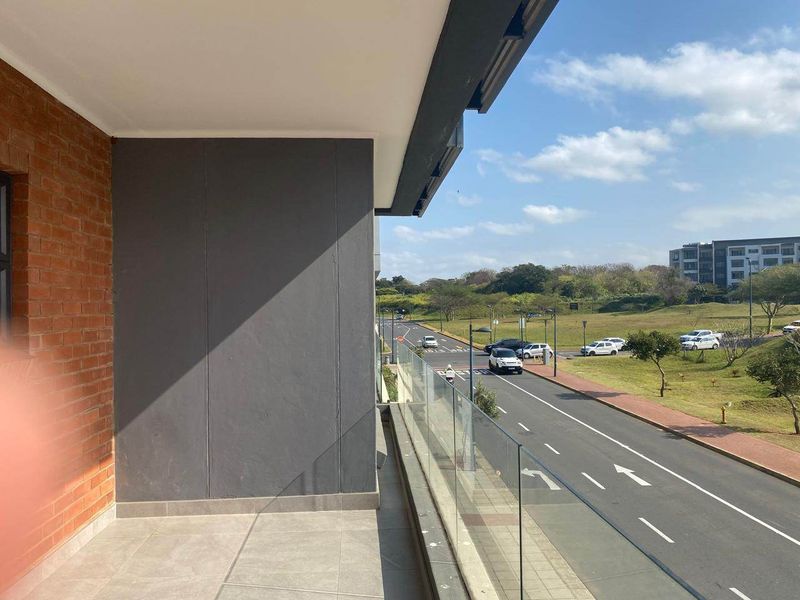A modern balcony featuring a glass balustrade, tiled flooring, and an exposed brick feature wall, offering an open view of the surrounding street and landscape.