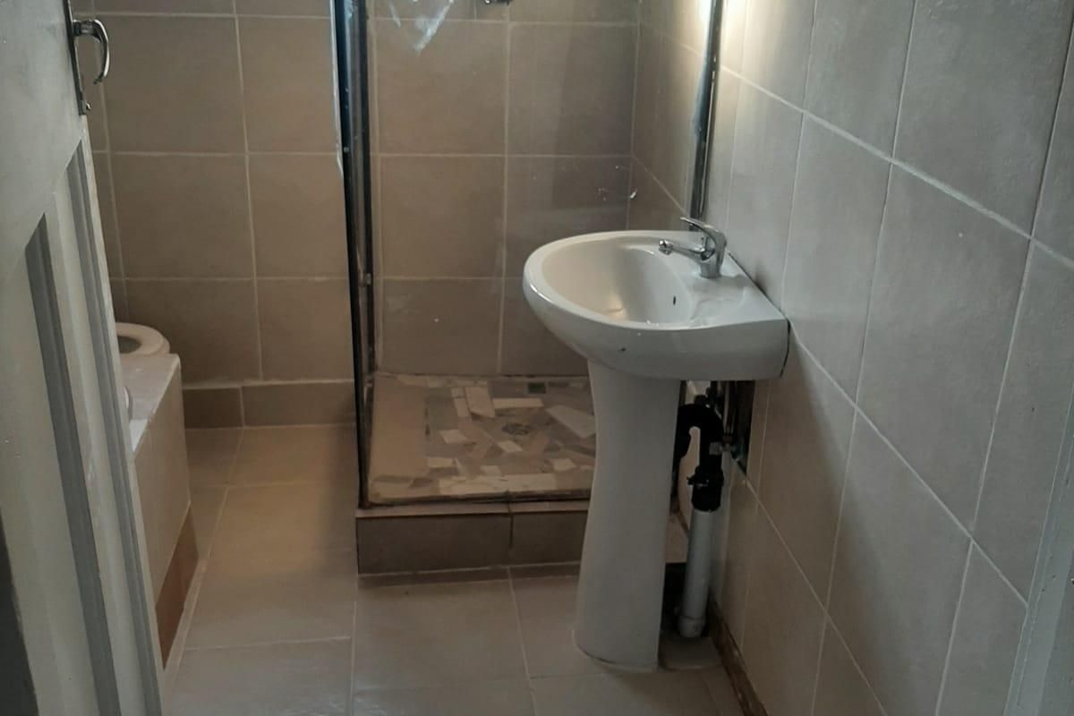 A compact bathroom featuring a pedestal basin, shower enclosure, and tiled finishes, currently requiring minor finishing work to the plumbing and wall edges.