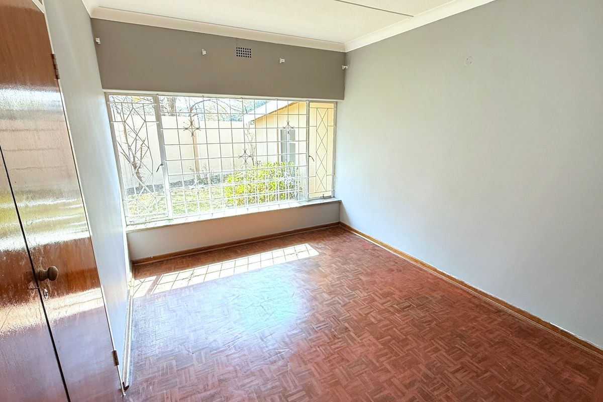 A bright, empty bedroom featuring classic parquet flooring, built-in wooden cupboards, and large windows fitted with security bars.
