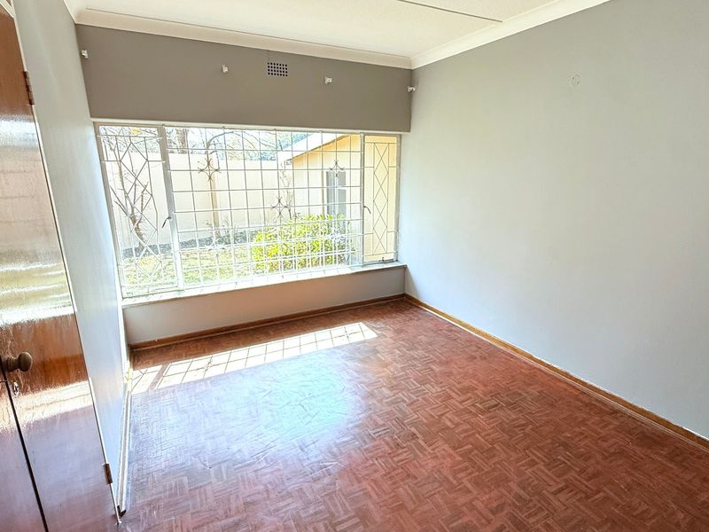 A bright, empty bedroom featuring classic parquet flooring, built-in wooden cupboards, and large windows fitted with security bars.