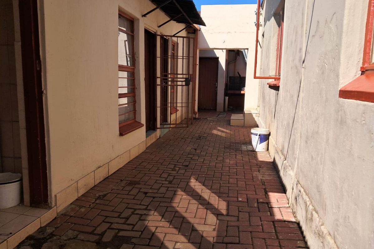 A narrow paved exterior walkway between building sections, featuring security gates and steel-framed windows, showing signs of wear and maintenance requirements.