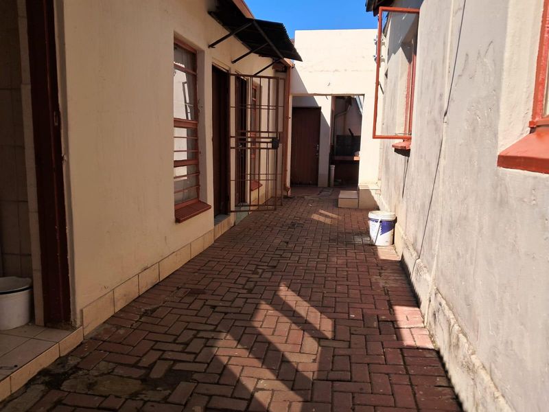 A narrow paved exterior walkway between building sections, featuring security gates and steel-framed windows, showing signs of wear and maintenance requirements.