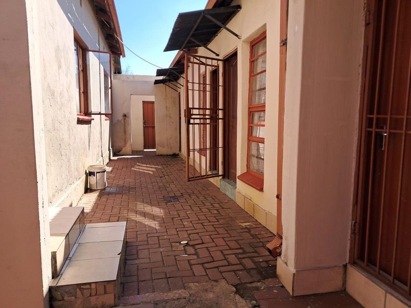 A narrow exterior rear walkway featuring brick paving, security gates, and metal window frames, providing access between building sections.