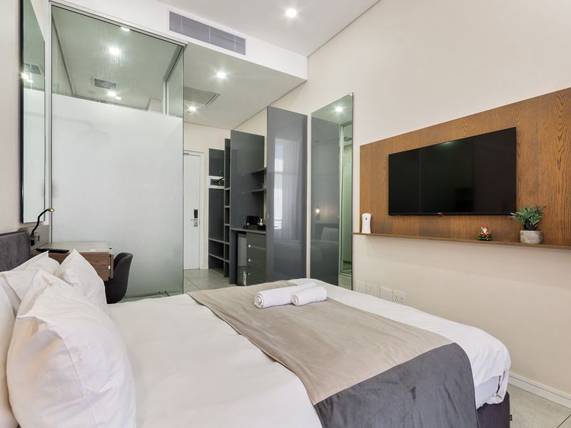 Modern bedroom featuring a glass-enclosed ensuite, built-in cabinetry, wood-paneled feature wall, and recessed lighting in a contemporary apartment setting.