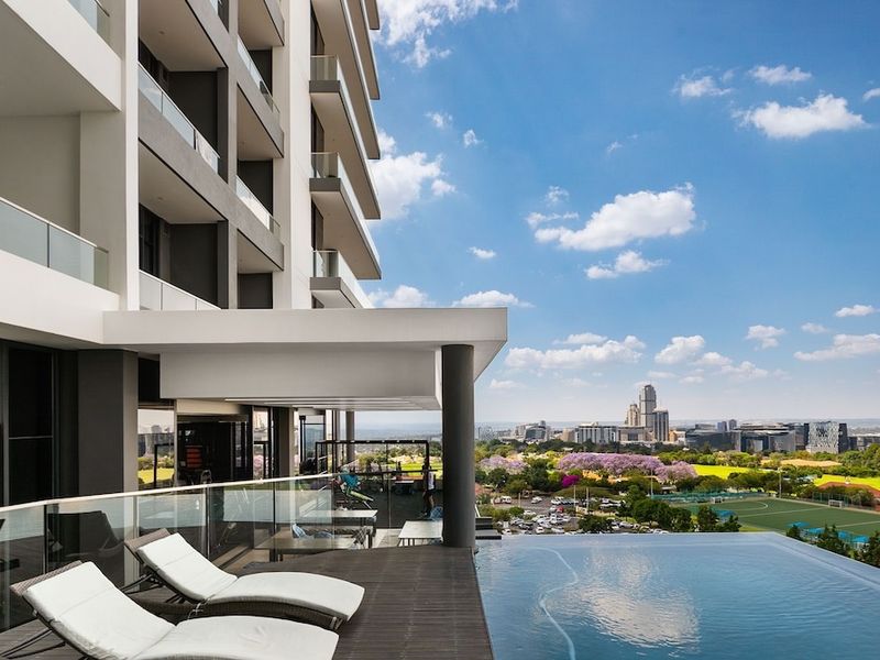 Modern apartment complex featuring an infinity pool with glass balustrades and a covered patio area overlooking the city skyline.