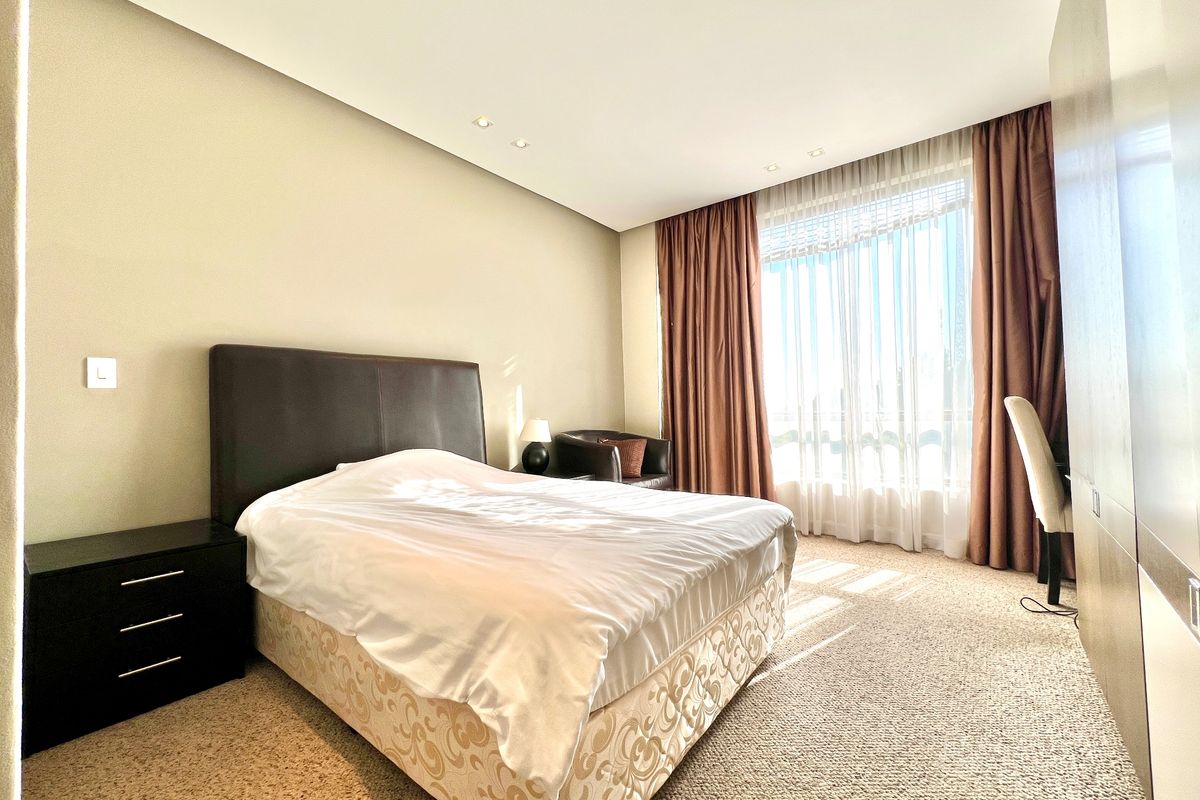 A well-lit bedroom featuring neutral walls, carpet flooring, recessed ceiling lighting, and a large built-in wardrobe with floor-to-ceiling curtains.