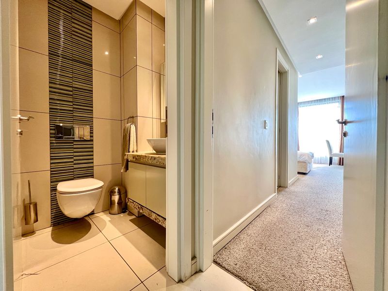 A modern bathroom featuring a wall-hung toilet, vanity with vessel sink, and decorative tiled wall accents, adjacent to a carpeted hallway.