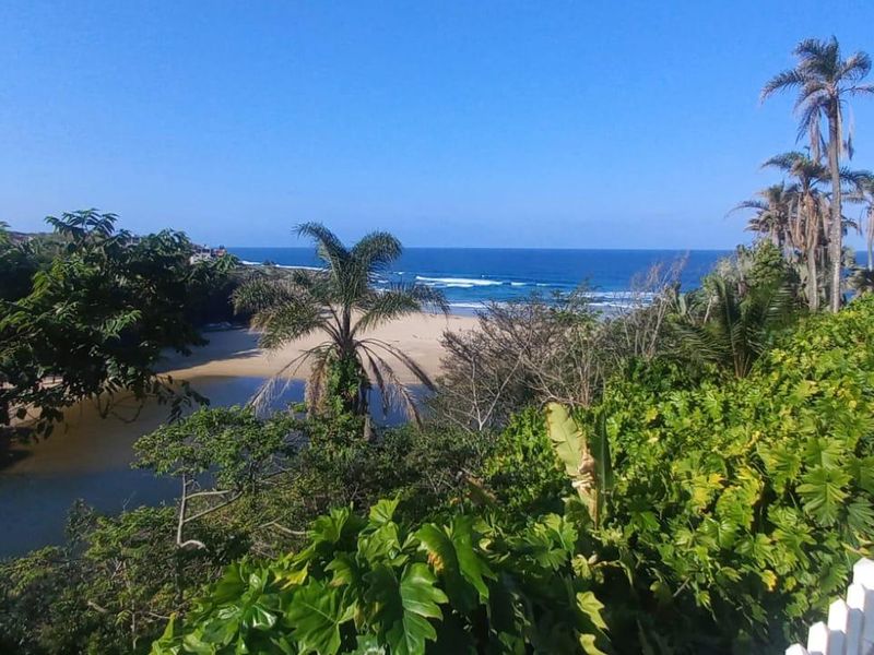 Scenic elevated view overlooking the coastline, beach, and estuary, framed by lush coastal vegetation and a white picket fence.