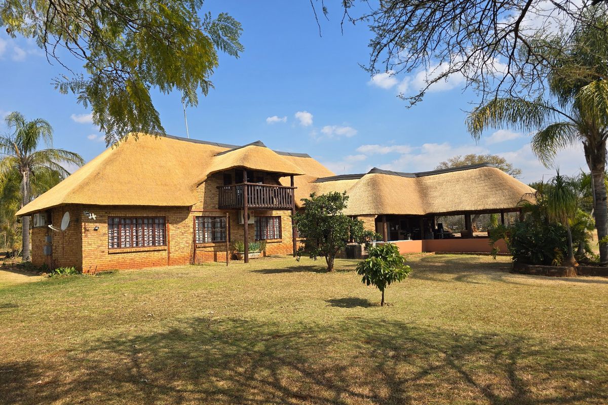 A spacious rear view of a traditional face brick house featuring a classic thatched roof, a timber balcony, and a large covered outdoor patio area.