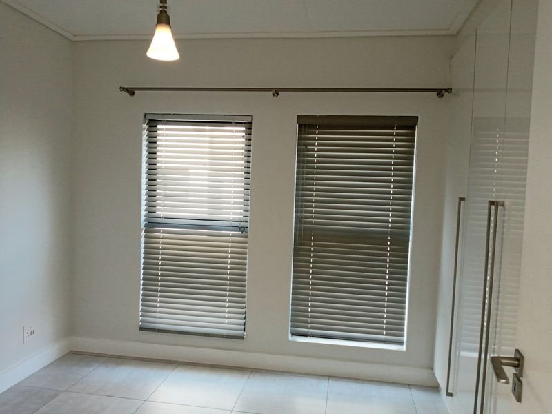 A clean, empty bedroom featuring neutral tiled flooring, built-in cupboards, and venetian blinds, offering a bright and versatile space.